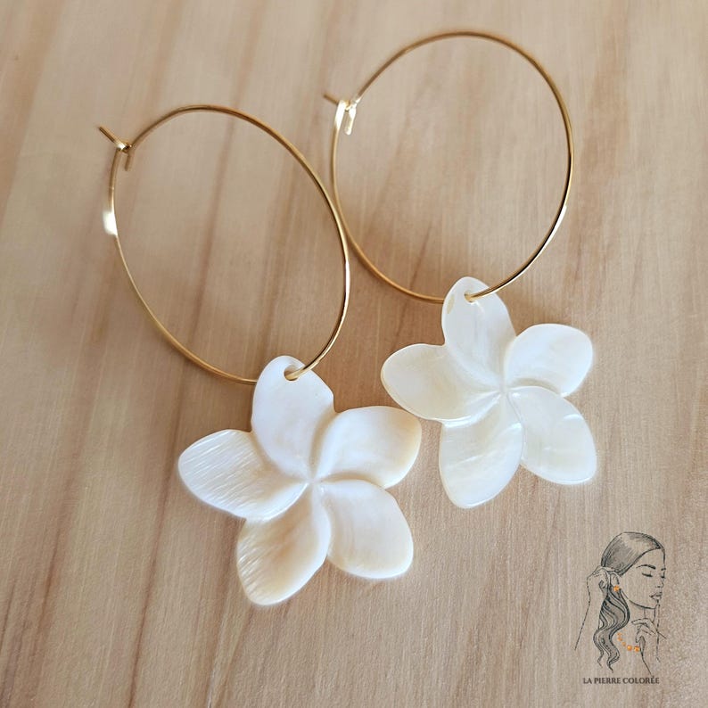 May include: A pair of gold hoop earrings with white, flower-shaped pendants. The earrings are set against a light brown wooden surface. The flower pendants have a pearlescent sheen and are detailed with petal lines.