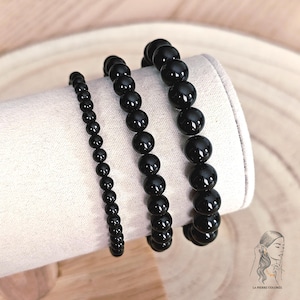 Op de afbeelding: Drie zwarte kralenarmbanden van verschillende maten worden getoond op een witte, cilindrische ondergrond. De kralen zijn rond en glanzend. De armbanden zijn waarschijnlijk gemaakt van onyx of een vergelijkbare steen. De achtergrond is licht en neutraal.