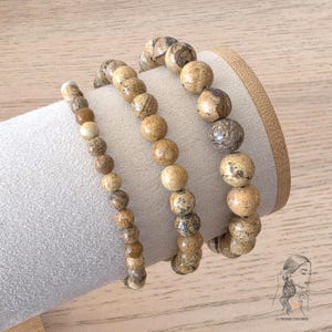 Puede incluir: Tres pulseras de cuentas con cuentas de piedra natural en tonos beige, marrón y crema. Las pulseras se exhiben en un soporte cubierto de tela beige. Las cuentas varían en tamaño y patrón.