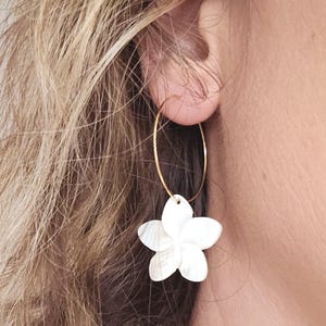 May include: A gold-colored hoop earring with a white, flower-shaped pendant. The pendant has five petals and is attached to the hoop. The earring is worn on a person's ear.