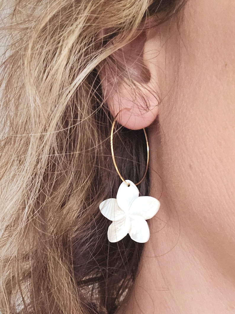 May include: A gold-colored hoop earring with a white, flower-shaped pendant. The pendant has five petals and is attached to the hoop. The earring is worn on a person's ear.