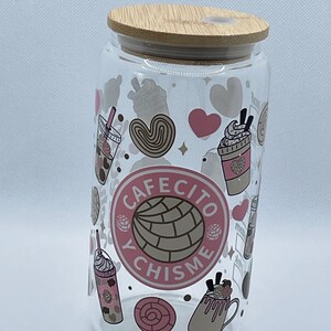 May include: A clear glass tumbler with a wooden lid, decorated with pink and brown illustrations of coffee drinks, pastries, and hearts. The tumbler has the words "Cafecito y Chisme" in a pink circle.