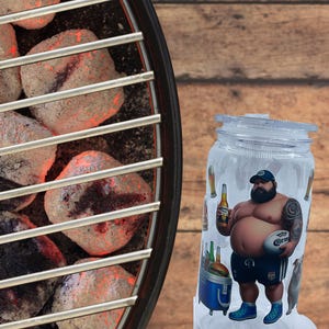 Może przedstawiać: Przezroczysty szklany słoik z pokrywką przedstawiający kreskówkową ilustrację mężczyzny trzymającego piwo i piłkę do rugby. Słoik ma motyw grilla, ze zdjęciami butelek piwa i lodówki. Tło to drewniana powierzchnia.