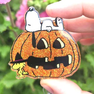 Puede incluir: Un portatarjetas de acr&iacute;lico con una calabaza de Halloween naranja brillante con una cara negra tallada. Snoopy descansa sobre la calabaza, mientras que Woodstock, con un peque&ntilde;o sombrero de bruja negro, est&aacute; de pie a la izquierda de la base.