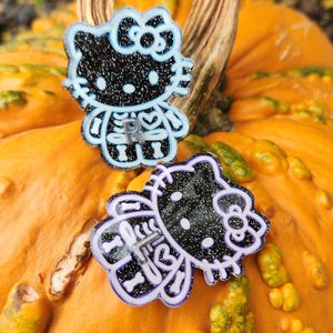 May include: Two Halloween-themed pins featuring a black and white glitter design of a cartoon cat with skeleton details. The pins are set against a bright orange pumpkin, creating a festive seasonal display.
