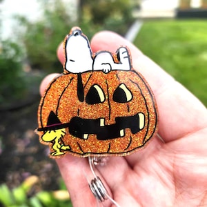 Puede incluir: Un portatarjetas de acr&iacute;lico naranja brillante con forma de calabaza tallada con cara negra. Snoopy est&aacute; acostado sobre la calabaza y Woodstock est&aacute; de pie a la izquierda con un peque&ntilde;o sombrero de bruja negro con una banda morada.