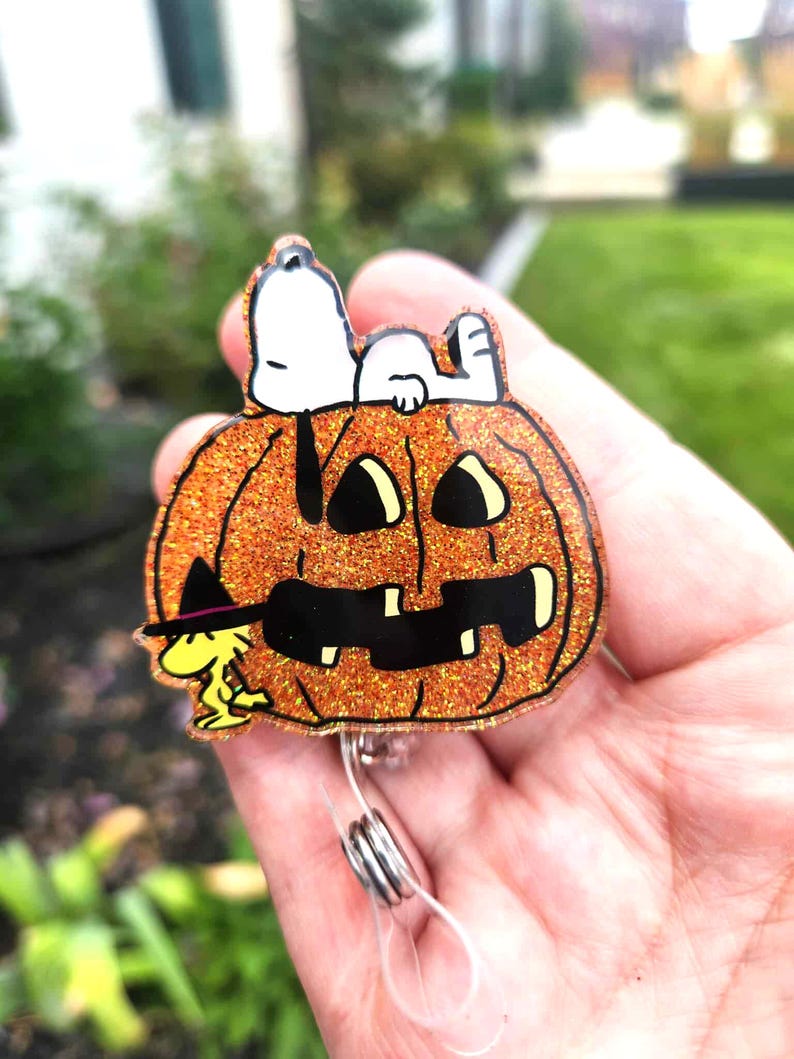 Puede incluir: Un portatarjetas de acr&iacute;lico naranja brillante con forma de calabaza tallada con cara negra. Snoopy est&aacute; acostado sobre la calabaza y Woodstock est&aacute; de pie a la izquierda con un peque&ntilde;o sombrero de bruja negro con una banda morada.