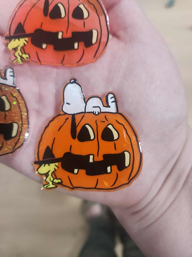 Puede incluir: Un broche de acr&iacute;lico brillante que muestra una calabaza de Halloween de color naranja intenso con una cara tallada en negro. Un perro blanco descansa sobre la calabaza, mientras un peque&ntilde;o p&aacute;jaro amarillo con sombrero de bruja est&aacute; al lado.