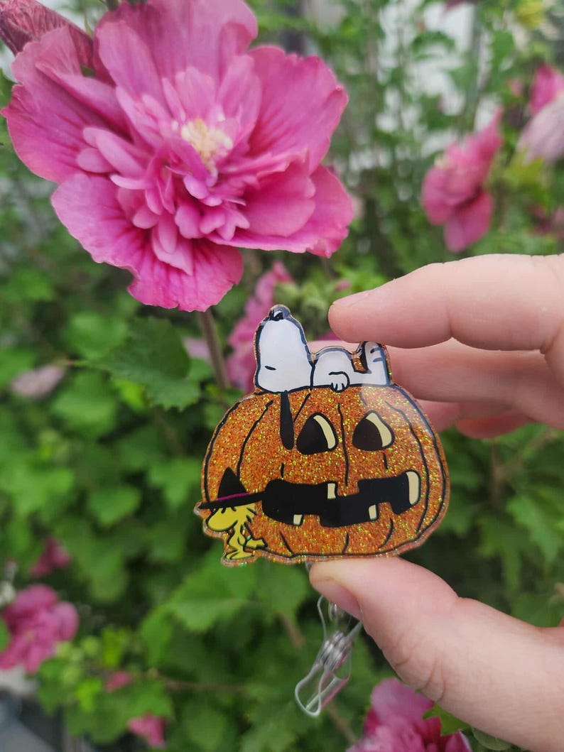 Puede incluir: Un portatarjetas de calabaza naranja con purpurina que presenta a un perro Snoopy blanco acostado encima y un p&aacute;jaro Woodstock amarillo con un sombrero de bruja negro en la base. El dise&ntilde;o tiene un acabado acr&iacute;lico transparente y un clip.