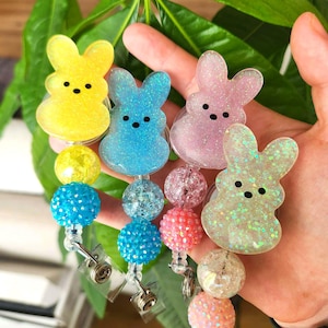 May include: A collection of colorful badge reels featuring glittery bunny-shaped toppers in yellow, blue, pink, and iridescent green. Each reel has a beaded accent and a retractable cord with a metal clip.