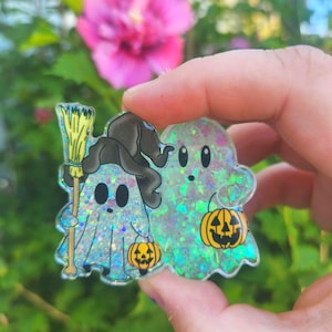 May include: A clear acrylic charm featuring two ghosts with iridescent glitter. One ghost wears a black witch hat and holds a yellow broom, while both ghosts carry small orange jack-o'-lantern buckets with black facial features and outlines.