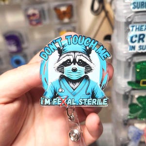 May include: A badge reel with a cartoon raccoon wearing a surgical mask and scrubs. The text on the badge reads "DON'T TOUCH ME" and "I'M FERAL STERILE". The badge is held by a clear plastic reel with a metal clip.