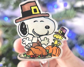 May include: A glittery acrylic badge reel featuring Snoopy and Woodstock wearing brown pilgrim hats. Snoopy stands behind a roasted turkey on a platter, while Woodstock sits on an orange pumpkin. The design includes black outlines and festive colors.