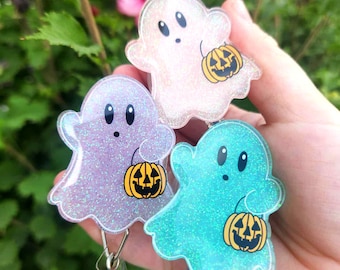 May include: Three glittery ghost-shaped badge reels in pink, purple, and teal. Each ghost holds a jack-o'-lantern and has black eyes. The badge reels have a clear plastic clip and a retractable cord. Perfect for Halloween.