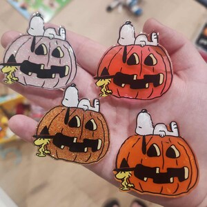 Puede incluir: Cuatro pines de acr&iacute;lico que muestran a Snoopy acostado sobre una calabaza de Halloween con Woodstock de pie a un lado. Las calabazas tienen varios acabados, incluyendo rosa claro con purpurina, naranja s&oacute;lido, naranja con purpurina y naranja transl&uacute;cido.