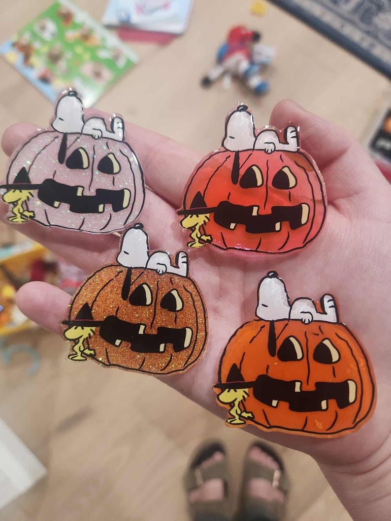 Puede incluir: Cuatro pines de acr&iacute;lico que muestran a Snoopy acostado sobre una calabaza de Halloween con Woodstock de pie a un lado. Las calabazas tienen varios acabados, incluyendo rosa claro con purpurina, naranja s&oacute;lido, naranja con purpurina y naranja transl&uacute;cido.