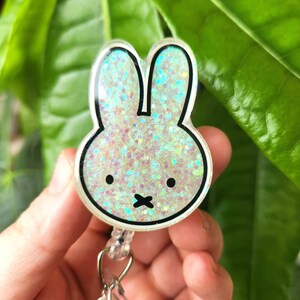 May include: A white and glittery bunny-shaped badge reel with a black outline and black eyes and mouth. The badge reel has a clear plastic retractable cord and a metal clip. The background is blurred green leaves.