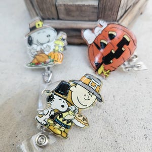 Puede incluir: Tres portatarjetas con personajes de Peanuts. Uno muestra a Snoopy con sombrero de peregrino y una calabaza, otro es una calabaza naranja tallada con Snoopy encima, y el tercero muestra a Charlie Brown y Snoopy con sombreros de peregrino.