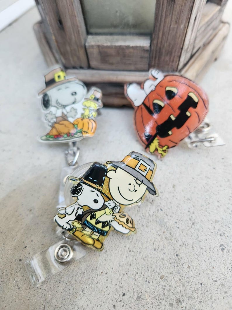 Puede incluir: Tres portatarjetas con personajes de Peanuts. Uno muestra a Snoopy con sombrero de peregrino y una calabaza, otro es una calabaza naranja tallada con Snoopy encima, y el tercero muestra a Charlie Brown y Snoopy con sombreros de peregrino.