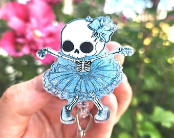 May include: A badge reel featuring a cartoon skeleton in a blue tutu and bow. The skeleton has a white skull and a blue tutu with glitter. The badge reel has a clear plastic retractable cord and a metal clip.