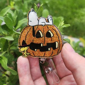 Puede incluir: Un portatarjetas de acr&iacute;lico naranja brillante con una calabaza tallada con una cara de jack-o'-lantern negra. Snoopy est&aacute; acostado sobre la calabaza, mientras que Woodstock, con un peque&ntilde;o sombrero de bruja negro, est&aacute; de pie a la izquierda.