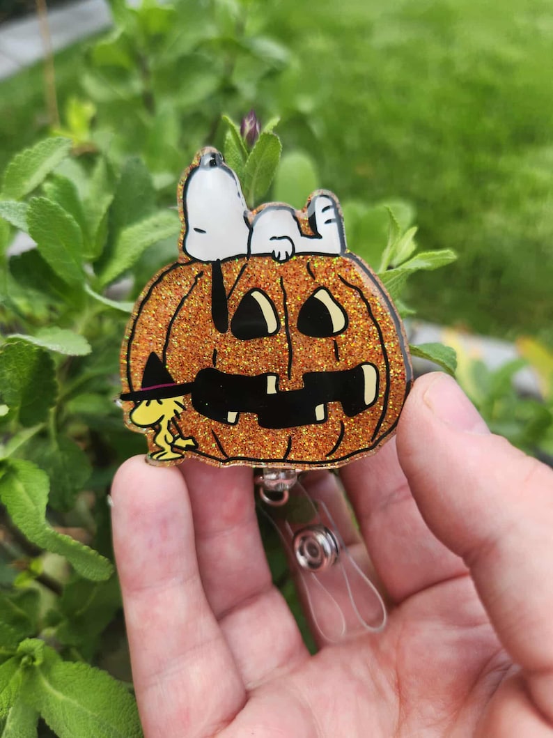 Puede incluir: Un portatarjetas de acr&iacute;lico naranja brillante con una calabaza tallada con una cara de jack-o'-lantern negra. Snoopy est&aacute; acostado sobre la calabaza, mientras que Woodstock, con un peque&ntilde;o sombrero de bruja negro, est&aacute; de pie a la izquierda.
