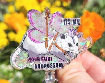 May include: A badge reel featuring a cartoon opossum with fairy wings, a crown, and a wand. The text on the badge reads "ITS ME" and "YOUR FAIRY GODPOSSUM". The background is blurred with yellow and orange flowers.