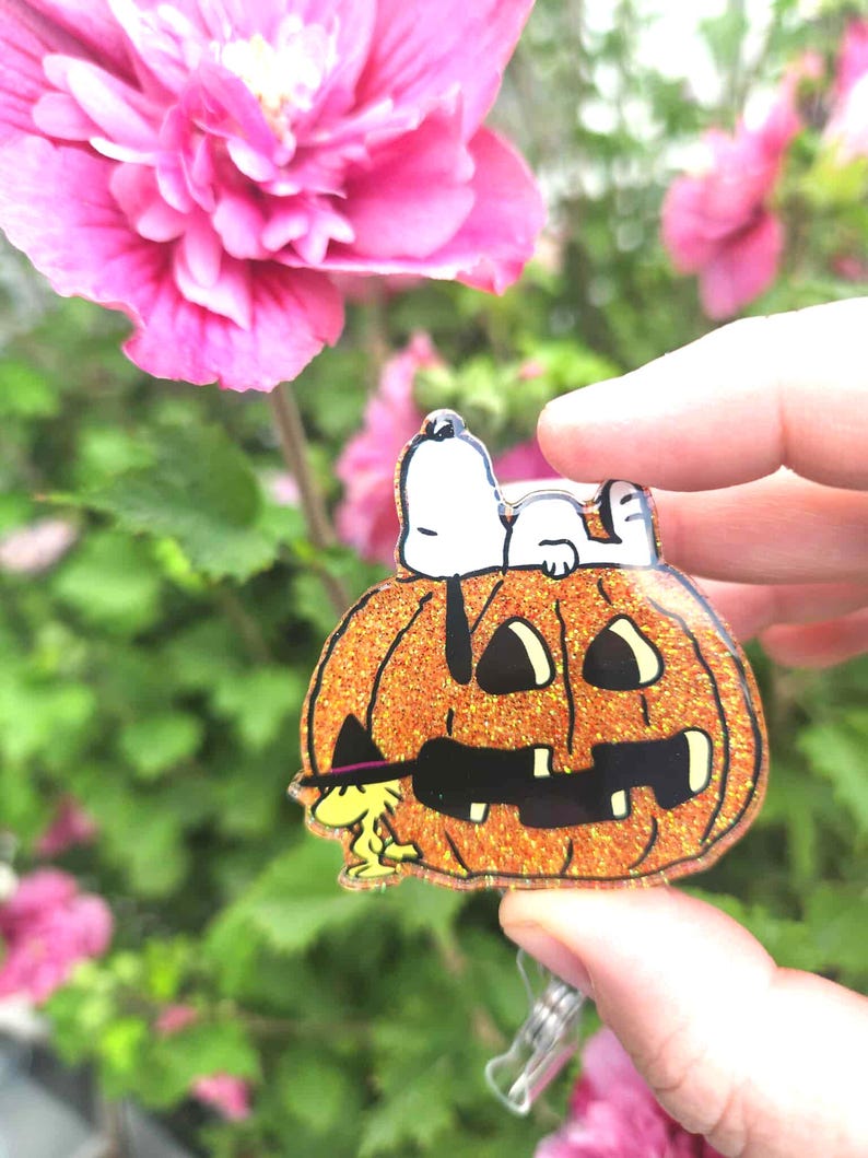 Puede incluir: Un portatarjetas de acr&iacute;lico con una calabaza de Halloween naranja brillante con una cara negra tallada. Snoopy descansa sobre la calabaza, mientras que Woodstock, con un peque&ntilde;o sombrero de bruja negro, est&aacute; de pie a la izquierda de la base.