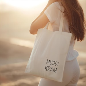 Könnte beinhalten: Eine cremefarbene Tragetasche mit dem schwarzen Aufdruck "MUDDI KRAM.". Die Tasche wird von einer Person in weißer Kleidung getragen. Der Hintergrund ist verschwommen und hat warme Farbtöne.