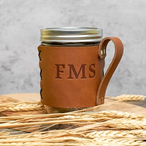 May include: A brown leather mason jar holder with a handle and the initials "FMS" embossed on the front. The holder is wrapped around a clear glass mason jar.