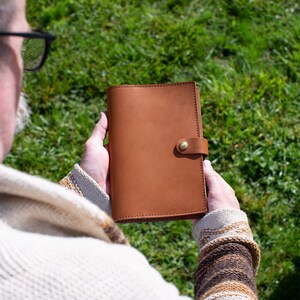 May include: A person holding a brown leather journal with a brass button closure. The journal has a stitched edge and a smooth, textured surface.