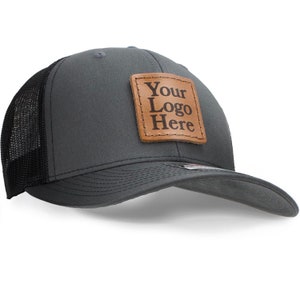 May include: A dark gray trucker hat with a black mesh back. The hat has a brown leather patch on the front with the text "Your Logo Here" in black.