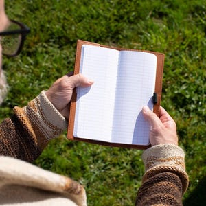 May include: Open notebook with lined pages held in a brown leather cover. The notebook is open, revealing blank pages ready for writing. The cover has a simple design with a black elastic closure. The background is a blurred green grassy area.