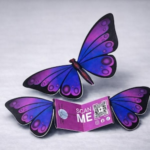 Könnte beinhalten: Zwei schmetterlingsförmige Objekte mit lila und blauen Flügeln und schwarzen Rändern. Die Flügel sind geöffnet und ein kleines lila Heft mit den Worten "SCAN ME" befindet sich zwischen den Flügeln. Das Heft hat auch einen QR-Code und Social-Media-Symbole.