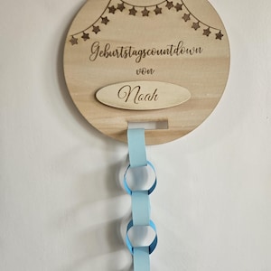 Könnte beinhalten: Holz-Geburtstags-Countdown-Tafel mit dem Text "Geburtstagscountdown von Noah" und einer hellblauen Papierkette. Die Tafel hat eine Schnur zum Aufhängen und eine Sternengirlande.