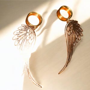 May include: A pair of gold-colored earrings featuring a wing design. The earrings have a circular top and a detailed wing pendant. The wings are a light copper color, with intricate feather detailing. The earrings are displayed on a light-colored surface.