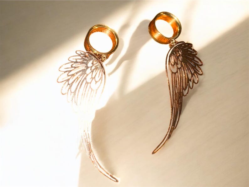 May include: A pair of gold-colored earrings featuring a wing design. The earrings have a circular top and a detailed wing pendant. The wings are a light copper color, with intricate feather detailing. The earrings are displayed on a light-colored surface.