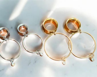 May include: Two pairs of ear gauges with dangling hoop attachments. One pair is silver-toned with clear round crystals, and the other pair is gold-toned with clear round crystals. Each gauge features a circular tunnel design.