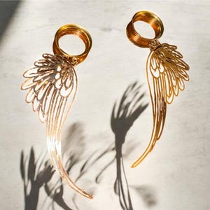 May include: Two gold-colored, wing-shaped earrings with intricate cut-out designs. Each earring features a circular top and a long, curved wing shape. The earrings cast shadows on a light-colored surface.