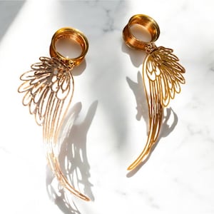May include: A pair of gold-colored ear gauges with a wing design. The gauges are made of metal and have a detailed, openwork pattern. The wings are long and slender, with a curved shape. The background is a white marble surface.