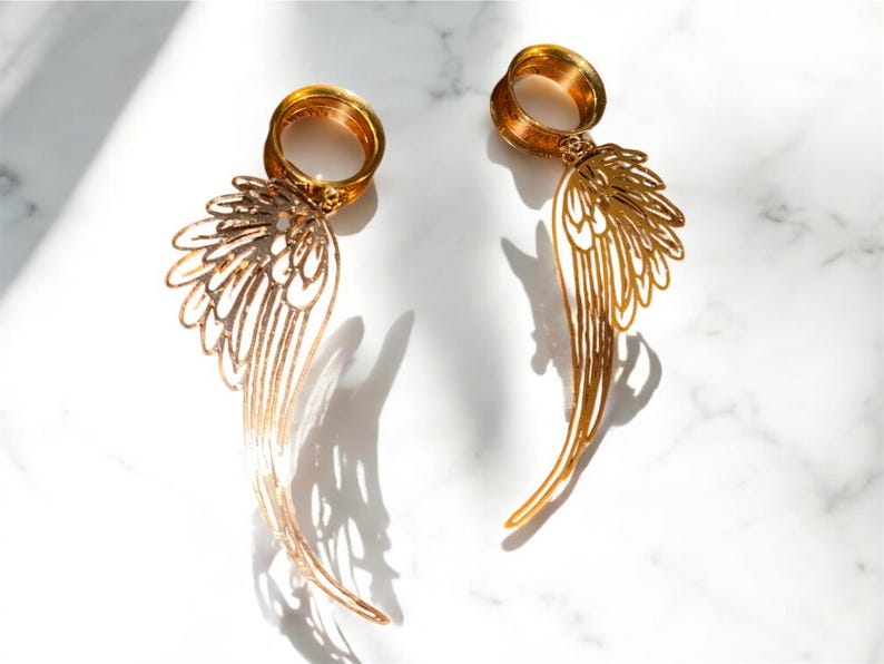 May include: A pair of gold-colored ear gauges with a wing design. The gauges are made of metal and have a detailed, openwork pattern. The wings are long and slender, with a curved shape. The background is a white marble surface.