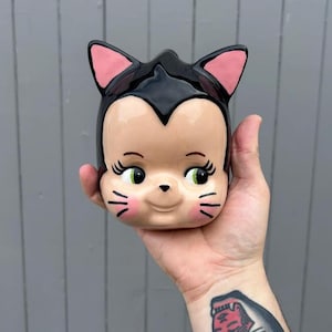 May include: A ceramic mug shaped like a cat head with a glossy black top, pink inner ears, and a beige face. The cat features large green eyes with long black eyelashes, a small black nose, and pink cheeks with three black whiskers on each side.