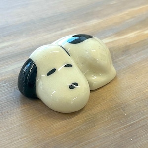 May include: A ceramic figurine of a cartoon dog, with a cream-coloured body and black accents on the ears and spots. The dog is lying down, with a glossy finish. The figurine is a decorative item.