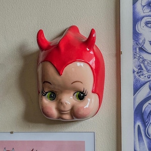 May include: A ceramic wall hanging of a baby devil face with red horns and a red head covering. The face has green eyes, long eyelashes, and rosy cheeks. The wall hanging is mounted on a light beige wall.