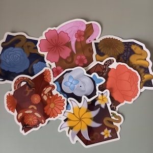 Op de afbeelding: Een verzameling gestanste stickers met bloemen- en slangenmotieven. De stickers tonen verschillende kleuren, waaronder roze, blauw, oranje en geel. De ontwerpen omvatten bloemen, slangen en decoratieve elementen.