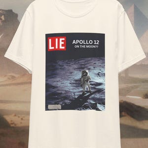 May include: White t-shirt with a graphic print of an astronaut on the moon. The graphic says "LIE APOLLO 12 ON THE MOON?"