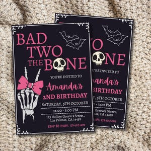 May include: Two black birthday invitations with pink text and white accents. The text reads "BAD TWO THE BONE" with a skeleton hand making a peace sign and a pink bow. The invitations are for Amanda's 2nd birthday.