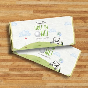 Pode incluir: Duas barras de chocolate embaladas com tema de golfe. As barras apresentam um design verde e branco com uma bola de golfe, um carrinho de golfe e o texto "Caleb's HOLE IN ONE! BIRTHDAY PAR-TEE". O fundo inclui nuvens e um campo de golfe.