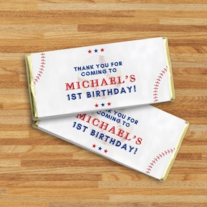 May include: Two white candy bar wrappers with baseball designs. The wrappers feature red stitching, a baseball graphic, and the text "THANK YOU FOR COMING TO MICHAEL'S 1ST BIRTHDAY!" on a wooden surface.