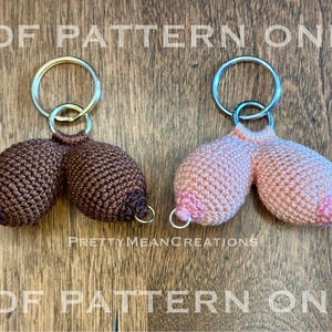 May include: Two crochet keychains, one brown and one pink, each shaped like a pair of breasts. The keychains have silver rings and are on a wooden surface. The text "PDF PATTERN ONLY" and "PrettyMeanCreations" are visible.